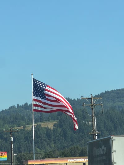 This is a landscape photograph taken in Woodland, United States of America, depicting an American flag waving against a clear blue sky. The flag is the primary subject, with its red and white stripes and blue field of stars prominently displayed. The flag is attached to a tall pole, which is partially obscured by a utility pole with power lines.
In the background, a dense, green forest covers a hillside, suggesting a rural or semi-rural setting. A large white truck or trailer is visible to the right of the flag, with some text on its side that appears to read "FlashCo" and "TRUCK ACCESSORIES." Below the truck, a vertical sign with colorful stripes and text is visible, with "PACIFIC P" being the most legible part.
The scene appears to be set during the daytime, under fair weather conditions. The angle of the photograph is slightly tilted, giving it a dynamic feel. The overall impression is one of patriotic display in a natural environment.