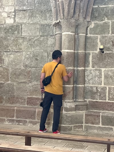 The image depicts a person standing inside a stone building, likely a church or abbey, in Le Mont-Saint-Michel, France. The person, viewed from behind, is wearing a yellow t-shirt, dark pants, and red and white sneakers. They have a black fanny pack slung across their chest and are holding a black camera in their right hand. Their left hand is resting on a stone pillar. The walls are constructed of large, rough-hewn stones, with some sections showing lighter-colored mortar. A simple wooden bench is visible in the foreground. A small candle holder is mounted on the wall to the right of the pillar. The overall environment appears to be dimly lit, with natural light possibly coming from an unseen source.