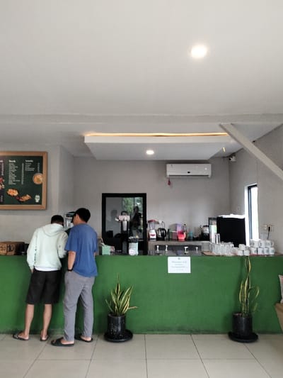 This image depicts an indoor scene at a casual self-service establishment, likely a cafe or restaurant, in Dramaga, Indonesia. Two men are standing with their backs to the camera at a prominent green counter. The man on the left wears a light-colored hoodie and dark shorts, while the man on the right is in a blue t-shirt and grey pants; both are wearing flip-flops. They appear to be ordering or waiting for items at the counter.
Behind the green counter, the background features a light grey wall equipped with an air conditioning unit and a security camera. Various items typically found in a service area are visible, including blenders, kettles, stacks of clean glasses, and white mugs. A dark-framed doorway or window in the back wall reflects a blurred figure. On the left wall, a dark menu board lists food and drink options, with headings like "Food" and "Drink," though specific items are mostly illegible.
On the green counter, a small white sign clearly states "Minuman self service" (Drink self-service) and "Terima Kasih" (Thank You), indicating a self-service beverage station. Two potted snake plants sit on the floor in front of the counter, adding natural decor. The ceiling is white with recessed lighting and a decorative dropped panel featuring strip lighting. The floor is light-colored tiled, and the space is brightly lit, suggesting it is daytime. The overall setting is functional and relaxed.