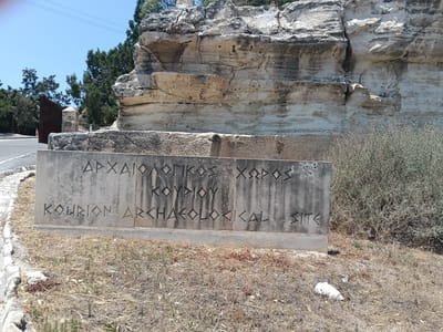 A rough-hewn concrete sign stands in a dry, grassy field under a clear blue sky. The sign has two lines of text carved into it. The top line is in Greek capital letters and reads "ΑΡΧΑΙΟΛΟΓΙΚΟΣ ΧΩΡΟΣ". The second line is in English capital letters and reads "KOURION ARCHAEOLOGICAL SITE". The sun is high in the sky, suggesting it is midday. Behind the sign, a large rock formation with distinct layers is visible, along with some dry brush. To the left of the sign, a paved road curves away, and beyond that, a tall, rusty metal gate and trees can be seen. The scene appears to be an entrance to an archaeological site, likely in a Mediterranean climate given the vegetation and rock formations. There are no people visible.