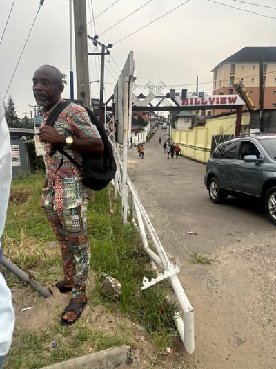 A man stands on a street corner near a fallen white sign, wearing a colorful shirt, trousers, and sandals. He has a black bag slung over his shoulder and a gold watch on his wrist. Behind him, the street leads to the "HILLVIEW" sign. A gray car is parked on the right side of the road, partially out of view, and there are other people walking in the distance. Overhead power lines are visible on the left, and the sky is overcast.