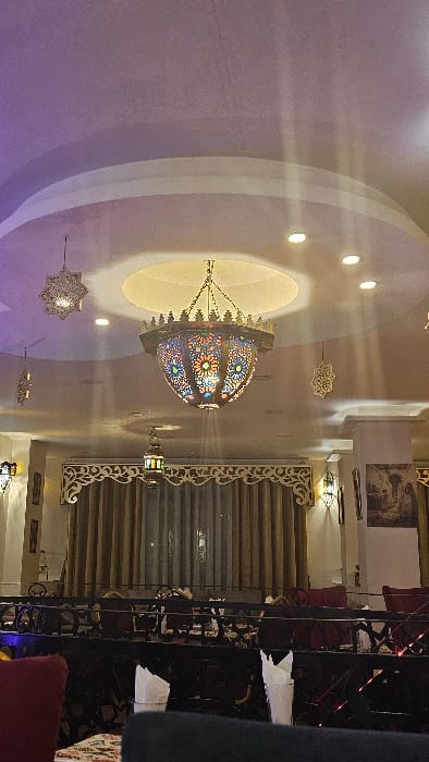 The photo shows an interior space with a large, colorful, domed chandelier hanging from a circular recess in a light purple ceiling. Decorative lighting fixtures and star-shaped ornaments adorn the ceiling. Ornate carved wooden panels and a set of blinds are visible on the right. An ornate, dark metal railing is present, alongside a glimpse of furniture.