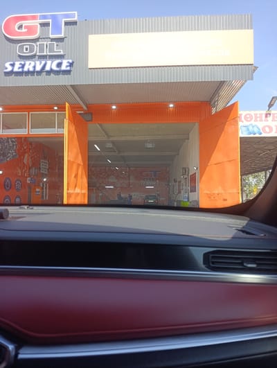 The image shows the entrance to a "GT OIL SERVICE" establishment, suggesting an automotive service center. The signage prominently displays "GT OIL SERVICE" in large, stylized letters. Above this, a smaller sign reads in Cyrillic, "СЕТЬ ПУНКТОВ ЗАМЕНЫ МАСЛА," which translates to "Network of Oil Change Points."

The service center has large orange doors, which appear to be open, revealing a spacious interior. The interior walls are decorated with graphics of car parts and possibly service-related imagery. Inside, a car can be seen in the distance. A few people are visible in the interior of the service bay. The scene appears to be set during daylight, with a clear blue sky visible.

The perspective of the photo is from inside a vehicle, looking out through the windshield. The dashboard of the car is visible in the foreground, with red upholstery. The image has a bright and clear quality, indicating fair weather. The location is identified as Ötegen batyr, Kazakhstan, which provides context for the Cyrillic text seen on the signage. The overall impression is that of a functional and modern auto service facility.
