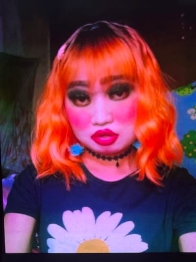 This media file is a close-up, tilted photograph of a person with bright orange hair and exaggerated makeup. The subject has large, dark eyes, pink blush on their cheeks, and bright red lips. They are wearing a black choker necklace with a small blue accessory attached. Their shirt is dark blue and features a large white daisy with a yellow center. The background is dark, with a hint of a colorful pattern, possibly a sheet or tapestry, behind the subject's head. The lighting appears to be artificial, casting a slight purplish hue on the scene. The image is blurry and appears to be taken on a low-resolution device, possibly a phone camera, with the subject looking directly at the lens. The orientation of the photograph suggests it might have been taken casually or spontaneously. No text is visible in the image. The overall impression is of a stylized or costume portrait, possibly for social media or personal expression. Given the location context of Cadek, Indonesia, it's possible this represents a cultural or personal aesthetic within that region, though no specific Indonesian cultural markers are explicitly identifiable.