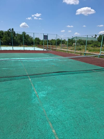 An outdoor tennis court with a green surface and a red border is shown. A net divides the court. In the background, there is another tennis court with a blue surface, fenced by a green metal mesh. A bicycle is parked next to the fence. Trees and a building with a green roof are visible beyond the fence. The sky is blue with scattered white clouds. The scene appears to be located in Gorkovskiy, Russia.