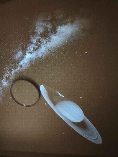 The image depicts a projected image of Saturn and a segment of the Milky Way galaxy on a brown, textured surface. Saturn, with its prominent rings, is positioned in the lower right of the frame, while the Milky Way appears as a luminous band of white specks in the upper left. The surface exhibits a granular texture, suggesting it could be a wall, ceiling, or floor. There are also several small, white dots scattered across the surface, resembling stars. A circular, reflective object, possibly a lens or a glass, is situated to the left of Saturn, near the Milky Way projection. The overall scene is dimly lit, with the projected images serving as the primary light sources. This image was captured in the city of Jiguan District, China.