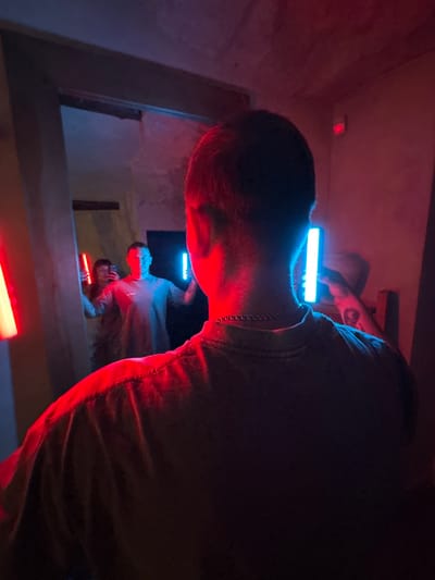 In the city of Postol, Russia, a person is standing in a dimly lit room, illuminated by red and blue neon lights. The subject is facing away from the camera, with their back to the viewer. They are holding a blue neon light stick in their right hand, which casts a bright blue glow on their face and neck. In the background, a mirror reflects another person holding a red neon light stick. The room appears to be a hallway or a narrow space, with textured walls. The atmosphere is dynamic and moody, with the contrasting colors of the lights creating a striking visual effect. The subject's attire is casual, and they are wearing a silver chain around their neck. The overall scene suggests a playful or artistic gathering, possibly at a nightclub or a private party.