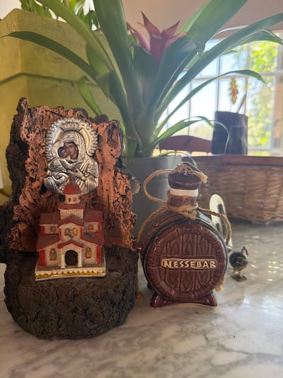 The image shows a still life arrangement on a marble surface. In the foreground, on the left, is a small religious figurine mounted on a dark, textured base. The figurine depicts the Virgin Mary holding the infant Jesus, rendered in silver, above a miniature church with a red roof and white walls. To the right of the religious figurine is a brown ceramic flask shaped like a barrel, with the word "NESSEBAR" inscribed on it. A twine rope is tied around the neck of the flask. A small, decorative globe on a stand is positioned to the right of the barrel flask. In the background, a large potted plant with green leaves and a pink flower is visible. Behind the plant, a window with natural light streaming through shows a blurred view of greenery and possibly furniture. The overall scene appears to be indoors, with the light suggesting daytime. There is no information in the image that specifies the location as Dromolaxia - Meneou Municipality, Cyprus.