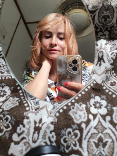 In a dimly lit room, a woman with auburn hair is captured in a mirror's reflection. She is holding a smartphone with a floral case in her right hand, her fingers adorned with bright red nail polish. Her left hand rests on her neck, and she is wearing a colorful, patterned blouse. The mirror reflects part of a patterned fabric, possibly a blanket or upholstery, in the foreground. In the background, the ceiling structure and a circular light fixture are visible. The scene suggests an indoor personal setting, possibly a home in Vytegra, Russia.