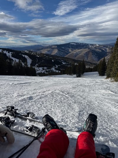 A first-person perspective shows a pair of skis and ski boots on a snowy slope. The person wears red ski pants. Ski poles are also visible. A mountain range is in the background with a blue sky and white clouds. Trees cover the mountain slopes. The snow on the ski slope appears granular and textured.