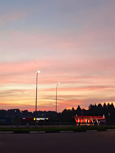 The image captures a twilight scene in Dubrovki, Russia, with a vibrant sky painted in hues of pink, orange, and blue. In the foreground, a paved road and a grassy median are visible, bordered by white and black concrete stoppers. Two lampposts stand tall on the median, their lights on, illuminating the darkening environment.

Further back, a row of evergreen trees forms a dark silhouette against the colorful sky. Below the trees, a brightly lit, modern gas station with red accents and white lights stands out. Adjacent to the gas station, a building with illuminated signage reading "МАГНИТ" (Magnit) is visible. In the distance, faint outlines of buildings, possibly with church spires, can be seen. The overall atmosphere is serene and tranquil, characteristic of a peaceful evening in a semi-urban or suburban setting.