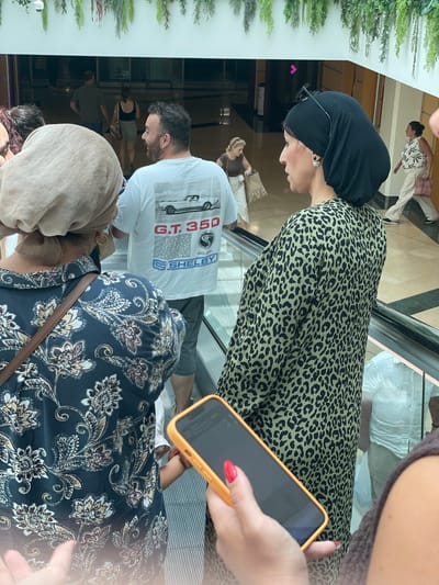 The image depicts a group of people on an escalator, likely in a shopping mall. The primary subject in the foreground is a woman wearing a black headscarf and a leopard-print dress, holding a yellow-cased smartphone. To her left, a man wearing a white t-shirt with a car graphic and the text "G.T. 350 SHELBY" is visible. Behind them, other individuals are seen on the escalator and in the background. The setting appears to be indoors with bright lighting, suggesting daytime. There are decorative green plants on the left side of the frame. The overall mood is casual and everyday. The location context provided is Algeciras, Spain.
