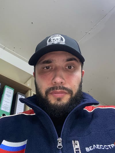 A medium shot captures a man from the chest up, looking directly at the camera. He is wearing a black baseball cap with a white logo featuring a skull and barbells. The man has a dark beard and a dark complexion. His jacket is navy blue with red and white accents on the collar and a white text on the right side of the chest. Behind him, to his left, is a shelf containing several folders, one of which is bright green and has visible text. The background appears to be an indoor setting with a light-colored ceiling and wall. The overall impression is a casual portrait, possibly taken in an office or personal workspace in Luzhitsy, Russia.