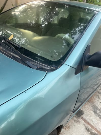 The image shows a close-up view of a light blue car, oriented sideways. The perspective is from the exterior, looking at the windshield and hood. The windshield is largely visible, reflecting the surrounding environment. Inside the car, obscured by the windshield's tint, are hints of seats and possibly a headrest, suggesting the interior of a vehicle. The hood of the car is a prominent feature, with a black antenna-like object mounted towards the top. The car appears to be parked outdoors, with a blurred background that hints at foliage and a wall, possibly indicating a residential or semi-urban setting. The lighting suggests it is daytime, but the specific time and weather conditions are not clearly discernible. The image captures a static scene, with no discernible activity. There is no visible text in the image. The overall impression is one of a parked vehicle in an outdoor environment.