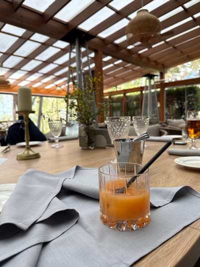 The image shows a table setting inside what appears to be a dining area with a sunroom-like structure, potentially a restaurant or a well-lit room in a building. A glass of orange liquid, possibly a cocktail, with a black straw stands prominently on a light grey napkin. A carafe of water and two glasses, a wine glass, and another beverage glass are visible further along the table. The table surface has a wood-grain texture. The sunroom structure is constructed with wooden beams forming a grid-like pattern, letting in natural light and providing views of greenery outside. The presence of a candlestick holder suggests a formal or upscale dining environment. While the location is specified as Baltezers, Latvia, the image itself doesn't intrinsically verify the location but it is an upper end restaurant.