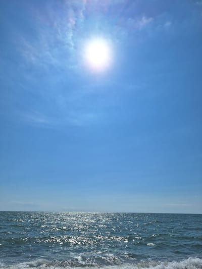 The image depicts a bright, sunny day at the sea in Kobuleti, Georgia. The sky is a vibrant blue with a few wispy clouds and the sun shining intensely, creating a lens flare effect. The sun's rays are reflected on the surface of the water, which is a deep blue with white caps on some of the waves. In the foreground, small waves are breaking on the shore, with white foam visible. The horizon line is clear, separating the sea from the sky. There are no people or man-made objects visible in the scene. The environment appears to be a natural coastal setting.