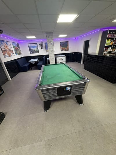 This image depicts the interior of a recreation room or bar located in Grau I Platja, Spain. The scene is devoid of people, active events, or interactions.
In the foreground and center, a regulation-size pool table with a green felt surface and a grey woodgrain-patterned exterior body stands on four black legs. A coin mechanism and display are visible on the front side of the table. The floor consists of large, light grey square tiles, displaying some minor scuff marks.
The walls feature a two-tone design: the lower half is covered with black marble-patterned tiles, while the upper half is plain white. A continuous purple LED strip light runs along the perimeter where the ceiling meets the upper walls. The ceiling is a suspended grid system with several recessed rectangular fluorescent light panels providing primary illumination.
On the left side of the room, a seating area includes two dark blue upholstered booth benches positioned opposite each other, with a small square table topped with a patterned white cloth between them. Above this, a black wall-mounted oscillating fan is installed. The wall in this section features several framed landscape and landmark posters, including one depicting a structure resembling the Taj Mahal, and others with mountain and natural scenery.
A white rectangular pillar with small square tiles stands towards the center-right, partially separating the seating area from other parts of the room. Adjacent to this pillar is a white chest freezer or cooler bearing the "TURIA" logo.
To the right, a black tiled bar counter extends along the wall, behind which a shelving unit displays various bottles of alcoholic beverages. A dark doorway is visible in the back right corner. Another dark doorway is partially visible on the far left.