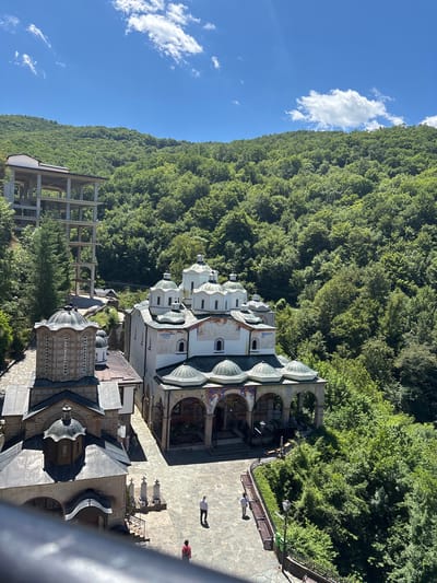 The image shows a complex of religious buildings, likely a monastery, nestled within a lush, green, mountainous landscape. The architecture features multiple white domed structures with intricate detailing, some adorned with colorful frescoes. In the foreground, a stone courtyard is visible with a few individuals walking. To the left, a partially constructed concrete building stands starkly against the natural backdrop. The sky is blue with scattered white clouds. The location is identified as Varovishte, North Macedonia.