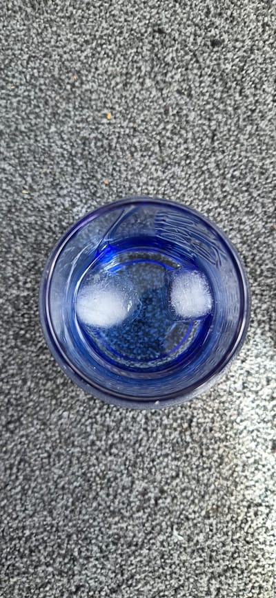 An overhead photograph captures a blue glass, filled with clear water and two irregular-shaped ice cubes, resting on a textured, speckled grey and white ground surface. The ground exhibits fine granular detail consistent with concrete or asphalt, interspersed with sparse, small orange and dark particles. The blue glass displays faint vertical and horizontal ridged patterns, visible both on its exterior and through the water, particularly near the rim. The water clearly reflects the blue interior of the glass. The ice cubes are distinct white solids with visible frosty textures, floating freely within the water. The scene is brightly lit by ambient light, suggesting an outdoor environment. This scene is located in Kolbermoor, Germany.