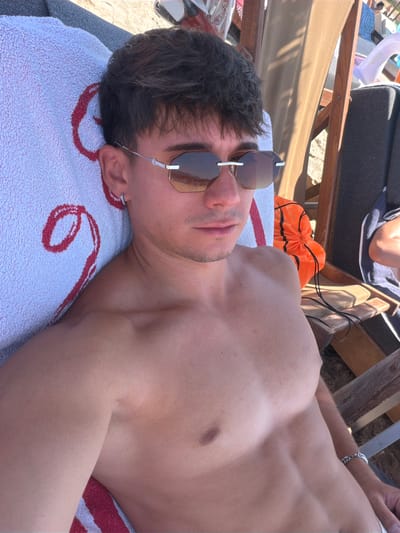 A young man with dark hair and sunglasses lies on a beach towel on a lounge chair at the beach. He is shirtless, wearing sunglasses, and has a silver earring in his left ear. The beach is sandy, and there are other lounge chairs and beachgoers visible in the background. The man is looking at the camera.