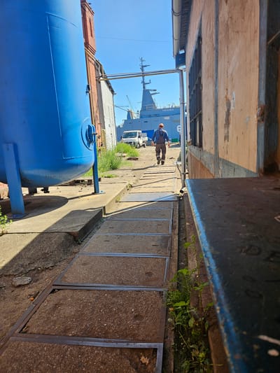 In Gdańsk, Poland, a man in a blue checkered shirt, brown pants, and an orange hard hat walks away from the camera down a concrete pathway. He is moving towards a large gray naval vessel docked in the background, which has the name "CZARKA" visible on its hull. To the left of the pathway is a large blue industrial tank. To the right, a weathered building with wooden siding partially obstructs the view of the ship. The scene appears to be an industrial or shipyard area, with a clear blue sky overhead.