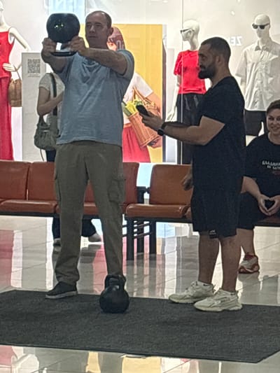 An indoor scene, likely a public space with tiled flooring and seating, features two men and several mannequins. In the foreground, a man wearing a blue t-shirt and khaki cargo pants is holding a black kettlebell with both hands in front of his chest. To his right, another man in a black t-shirt and black shorts stands, looking to his left and holding a smartphone. A woman is partially visible to the far right, seated and wearing a black t-shirt with Russian text. In the background, mannequins are displayed, some wearing clothing. The setting appears to be a commercial or retail environment, possibly a mall, given the mannequins and seating area. The presence of a kettlebell suggests a fitness-related activity or demonstration. The location is identified as Tri Protoka, Russia.