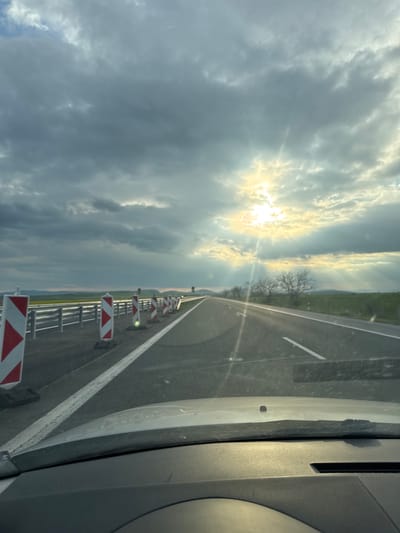 The image shows a view from inside a vehicle, possibly a car, traveling on a highway. The road has a white line marking the lane. Alongside the road, there are construction barriers with red and white striped markings. The sky is overcast with clouds and a bright spot indicating the sun. The landscape in the distance appears to be flat with some trees visible. There is no indication in the image that reveals that this scene is in the city of Chokoba, Bulgaria.