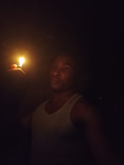A person, likely male, is holding a lit lighter in their left hand, which is positioned in the foreground of the image. The flame from the lighter illuminates the person's face and upper body. The person is wearing a light-colored tank top. The background is predominantly dark, suggesting a nighttime setting. The scene appears to be in a dimly lit outdoor environment, possibly within the city of Orodo, Nigeria. No other individuals or objects are clearly discernible in the image due to the low light conditions.