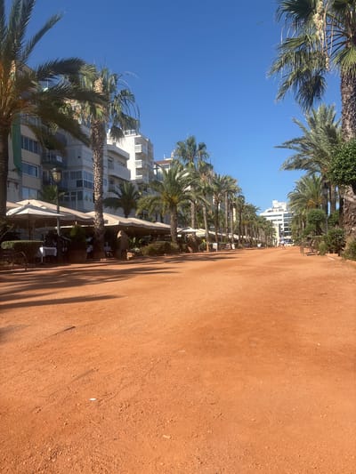 This is a photograph of a wide, reddish-brown dirt path lined with palm trees on either side, under a clear blue sky. In the background, there are several multi-story buildings with balconies and awnings. On the left side of the path, there are outdoor dining tables set up with white tablecloths and umbrellas. On the right side, there are more palm trees and some low-lying greenery. In the distance, a few people can be seen walking along the path. The scene appears to be a promenade or walkway in a coastal city, likely a tourist area. Based on the architectural style of the buildings and the abundance of palm trees, this location is consistent with the appearance of the promenade in Lloret de Mar, Spain.
