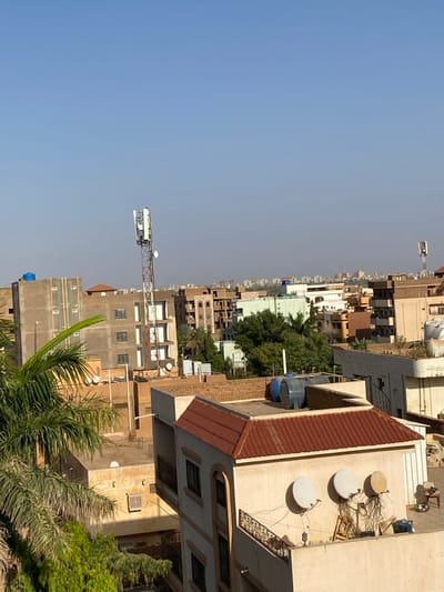 The image shows an overview of a residential area in Omdurman, Sudan. It's taken from a rooftop, looking towards the west, with a clear blue sky in the background. The image captures a dense urban landscape with multi-story buildings constructed from pale yellow and cream-colored concrete. There are several palm trees growing within the compound, and the rooftops are covered with satellite dishes and water tanks. There are multiple antennae towers visible in the distance, likely for communication purposes. The scene is predominantly made up of architectural elements, and there are no people visible.