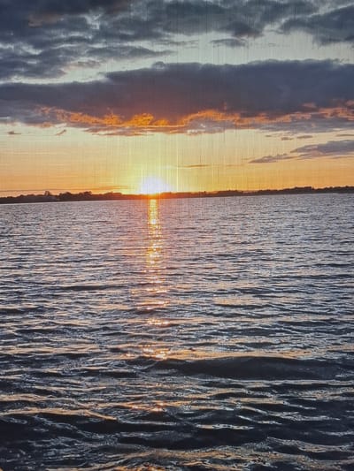 A wide shot captures a serene sunset over a calm body of water. The sky is painted with hues of orange and yellow, with scattered dark clouds adding depth and contrast. A faint silhouette of a treeline can be seen on the horizon, separating the sky from the water. The water's surface is gently rippled, reflecting the warm glow of the setting sun. The scene appears to be a natural landscape, likely a lake or a calm sea, and there are no people or man-made structures immediately visible. The image's resolution suggests it might have been taken with a camera or phone that has a visible screen or grid overlay. The location is described as Tecnimont camp, Russia.