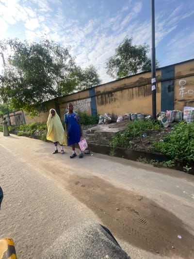 In Akowonjo, Nigeria, two young girls, one wearing a yellow hijab and the other a blue school uniform, stand on the side of a paved road. The girl in the hijab stands slightly behind the girl in the blue uniform, who is holding a pink tote bag. Behind them, a weathered, yellow and blue striped wall runs along the road, with overgrown vegetation in front of it. Several large, burlap sacks are piled along the base of the wall. A tall, dark lamppost stands near the wall, with stickers attached to it. In the background, trees with dense foliage are visible against a sky with scattered clouds. The foreground of the image shows a portion of the paved road and what appears to be the leg of a person, suggesting the image was taken from their perspective.
