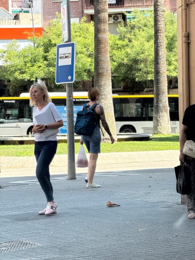 A street scene in L'Hospitalet de Llobregat, Spain, captures several individuals on a sunny day. In the foreground, a woman with blonde hair and glasses, wearing a white t-shirt and dark jeans, stands with a white folder in her arms. To her right and slightly behind, another woman with short brown hair and visible arm tattoos, wearing a dark patterned tank top and denim shorts, walks with a transparent plastic bag in her right hand. Further to the right, the lower half of another person is visible, wearing dark patterned trousers and a light-colored shoulder bag. A bus, identified by the "moventis" logo, is visible in the background, alongside a bus stop sign displaying a bus icon. Palm trees and leafy green trees provide a backdrop, with buildings and an air conditioning unit visible in the distance. The ground is paved with grey interlocking bricks.