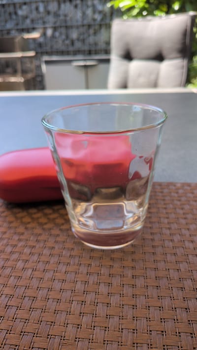 A clear glass tumbler sits on a brown woven placemat. The glass is partially filled with a clear liquid, reflecting the red object behind it. The red object appears to be a bottle with a rounded bottom. In the background, out of focus, is a patio setting with green foliage to the left and a grey tiled surface and light-colored furniture to the left. The overall impression is a casual outdoor scene, possibly on a patio or balcony. The lighting suggests daytime, with soft shadows. No text is visible in the image.