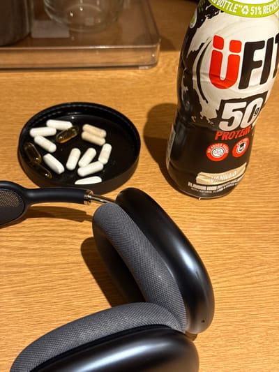 The image depicts a still life scene on a light-colored wooden surface, possibly a table or countertop, in an indoor setting. In the foreground, a pair of black over-ear headphones, resembling Apple AirPods Max, are partially visible, featuring grey mesh earpads and a black metallic exterior. To the left of the headphones, a black circular lid, likely from a container, holds approximately ten white opaque capsules, four translucent amber-colored capsules, and three light brown opaque capsules. To the right, a black uFit 50g Protein shake bottle with "Vanilla" flavor is standing upright. The bottle features white and red branding, including "NO ADDED SUGAR" and "LOW FAT" labels. The background is blurred, showing indistinct shapes including what appears to be a clear glass container and a metallic object. Shadows are cast from the left, suggesting a light source in that direction. The specific location within Mid Suffolk, United Kingdom, cannot be determined from the visual evidence provided in the image.