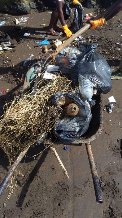 The image shows a wheelbarrow filled with trash on a dirt path in Limbe, Cameroon. The wheelbarrow contains dry grass, multiple plastic bags, and assorted plastic bottles and other debris. A person is visible in the background, holding a wooden stick and wearing a black shirt and yellow gloves. They are likely involved in collecting and organizing the trash. The scene is in a natural setting, possibly near a beach or riverbank. The environment is characterized by dirt and scattered debris, indicating a lack of organized waste management. The overall impression is of a community struggling with waste disposal and a need for proper sanitation.