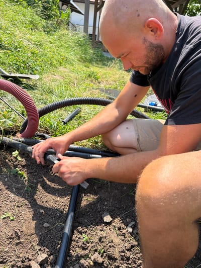 In Bašť, Czechia, on a sunny day, a bald man with a beard is kneeling on the ground, wearing a black t-shirt and beige shorts. He is focused on working with black plastic pipes that are partially buried in the soil. His hands are holding a tool, possibly a pipe cutter, and he appears to be in the process of cutting or attaching one of the pipes. Nearby, a red corrugated hose and another black corrugated hose are visible, lying on the grass and dirt. The environment is natural, with green grass and some vegetation, suggesting an outdoor setting, possibly a garden or a construction site.