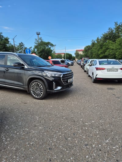 A row of cars is parked along a paved road, lined with green trees and a clear blue sky overhead. The closest car in the foreground is a black SUV with chrome accents and distinctive silver multi-spoke wheels. Beyond it, a line of other vehicles, including a white Hyundai Accent with a license plate reading "KZ 644 AKF 14", are parked. In the background, buildings with red roofs and utility poles are visible amongst the trees. Traffic signs, including a blue circular sign with a white arrow pointing left and an octagonal red stop sign, are present. The scene appears to be in a city, possibly near an administration building, given the mention of Aqsu city administration. The sunlight suggests it is daytime, and the clear sky indicates fair weather. No people are visible in the image.