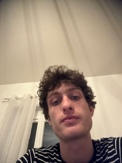 A young adult male, characterized by dark curly hair, light-colored eyes, and a neutral expression, is depicted in a close-up, head-and-shoulders shot, looking directly at the camera. He is wearing a dark t-shirt with light horizontal stripes. The scene is an indoor residential setting in the city of Roanne, France. The background consists of a plain, light-colored ceiling or upper wall, exhibiting multiple faint, parallel, vertical shadow lines. To the left, a window with a white frame is partially visible, covered by a sheer white curtain suspended from a silver-toned rod via ring eyelets. The ambient lighting appears to be artificial.