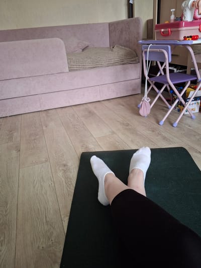 The image shows a room interior. In the upper left corner, there is a pink toy playset and a light purple folding chair. Next to them is a light pink sofa with a textured pillow. A dark green yoga mat is placed on a light brown wooden floor. A person wearing black leggings and white socks is lying on the yoga mat. The location in the city of Ozyorny, Russia, cannot be determined from this image.