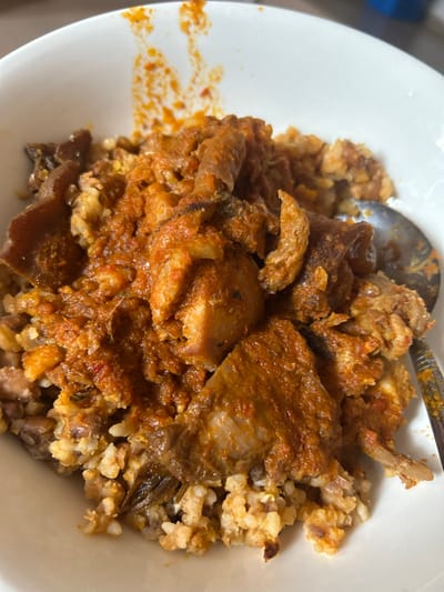 A white ceramic bowl, partially stained with reddish-orange residue on its interior rim, is centered in the frame. Inside, a serving of food consists of a base of light brown and off-white grains, likely rice and beans, mixed with darker fragments. This base is generously covered with a thick, reddish-orange stew or sauce. Within the stew are multiple pieces of what appear to be cooked meat or possibly fish, along with several pieces of cooked cow skin (ponmo), identifiable by their distinct texture and darker coloration. A metallic spoon, with some reddish residue on its handle, is partially visible along the right edge of the bowl, resting within the food. The scene depicts a meal prepared and consumed in Egbe, Nigeria. Natural light illuminates the dish from the upper left, casting subtle shadows within the food. No individuals are present in the image.