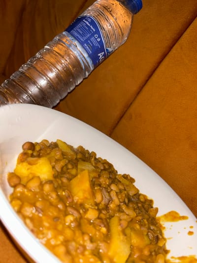 A water bottle labeled "Aqua" with a blue cap sits beside a white plate of cooked lentils and potatoes in a yellow curry sauce. The objects are set against a plush, orange surface, possibly a sofa or cushion. The water bottle is covered in condensation droplets. The food appears to be a hearty, home-cooked meal, likely a vegetarian dish.