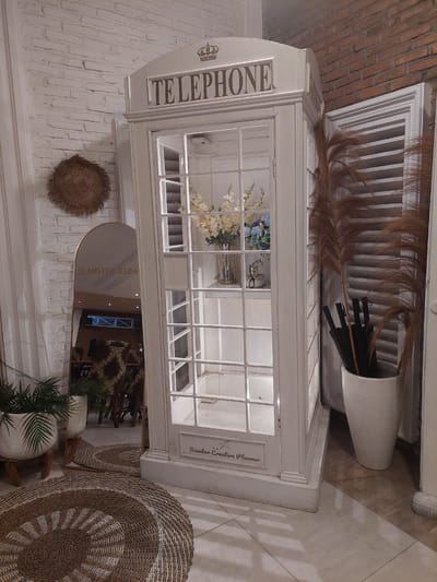 This media file depicts a quaint, decorative telephone booth, painted white and filled with artificial flowers and decorative grass. The booth is a prominent object within what appears to be an indoor setting with white brick walls and tiled flooring. Several other decorative objects are visible, including a woven circular mat on the floor and a mirror on the wall reflecting the interior of the space, which hints at a restaurant or cafe environment with patterned chairs and tables. The overall aesthetic is rustic and charming, with a focus on natural and handmade elements.
The telephone booth itself has the word "TELEPHONE" emblazoned on its side, along with a crown emblem. There is also text on a small plaque attached to the side of the booth that reads "Sianlan Creative Planner". The mirror on the wall displays the text "MISTER KEBA" with an Instagram-like camera icon, suggesting a business name or social media handle. The lighting in the scene suggests it is daytime or a well-lit interior space. There are no people visible in the image, and the activity seems to be a static display rather than a dynamic event. The overall mood is calm and inviting, suggesting a place designed for aesthetic appeal and perhaps relaxation. The location context of Pematang Siantar, Indonesia, adds a specific geographical detail to the scene.