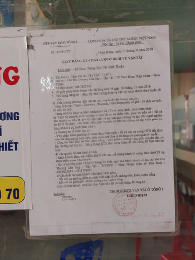 A framed document titled “GIẤY ĐĂNG KÝ CHẤT LƯỢNG DỊCH VỤ VẬN TẢI” (QUALITY REGISTRATION OF TRANSPORTATION SERVICES) is affixed to a surface. The document is addressed to Sở Giao Thông Vận Tải Ninh Thuận (Department of Transport of Ninh Thuận). The document provides details about Hợp Tác Xã Vận Tải Ô Tô Số 1 (Transportation Cooperative No. 1), including the location at 126 Hải Thượng Lãn Ông - Tấn Tài - TP Phan Rang Tháp Chàm, Ninh Thuận; phone number 068.3825847; transport license; vehicle quality; facilities; operation; customer rights; customer information, phone numbers, and services for passengers. The document has a red stamp and signature from Nguyễn Hoàng Anh.