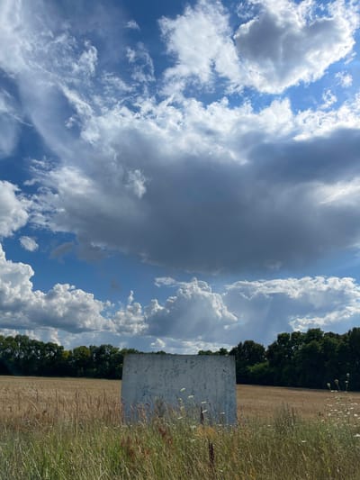 An outdoor scene captures a weathered, light blue rectangular structure situated in a dry, golden field. The structure, which appears to be some form of partition or panel, is positioned in the foreground and is partially obscured by tall, dry grass and some white wildflowers. Behind the structure, the field extends to a line of dense, dark green trees that form the horizon. The sky above is a vibrant blue, dotted with a variety of clouds. Some clouds are bright white and puffy, characteristic of cumulus, while others are larger, darker, and more diffused, suggesting the presence of storm clouds or heavy overcast. The light suggests it is daytime, with the sun casting shadows and illuminating the clouds. The location is identified as Stremyhorod, Ukraine.