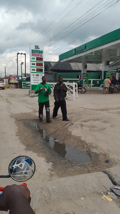 In Agbarho, Nigeria, two young men are walking across a paved area in front of a Bimbo Petroleum Ltd. gas station. The man on the left wears a green t-shirt and black pants, while the man on the right is dressed in a dark jacket and black pants. Both appear to be talking to each other. The gas station features a prominent digital sign displaying fuel prices, and "BIMBO PETROLEUM LTD" is visible on the canopy. Several motorcycles are parked at the station, with a few people visible in the background near the pumps. The ground in the foreground is uneven and damp, with a large puddle of muddy water. A motorcycle side mirror, partially broken, is visible in the lower left corner of the frame. The sky is overcast with dramatic clouds. The environment suggests a typical street scene in a Nigerian town.