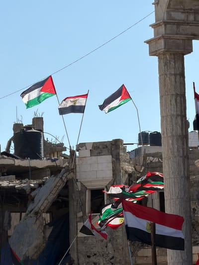 In the city of Jabalia, Palestinian Territories, the image depicts a scene of destruction and protest. In the foreground, several flags are prominently displayed. The flag of Palestine, with its black, white, and green horizontal stripes and red triangle, is seen waving. Adjacent to it, the flag of Egypt, featuring red, white, and black horizontal stripes with the Eagle of Saladin emblem in the center, is also visible. A cluster of flags, predominantly showing the colors red, white, black, and green, are seen further back, partially obscured by the debris.
The background reveals the aftermath of conflict, with severely damaged buildings. Concrete rubble, twisted metal reinforcements, and broken brick walls are evident, indicating significant structural damage. In the mid-ground, large black water tanks are mounted atop one of the damaged structures. A stone column, possibly part of a larger architectural element, stands tall on the right side of the image, contrasting with the surrounding destruction. The sky above is clear and blue. The scene suggests a context of ongoing conflict and resistance, with the flags serving as symbols of national identity and solidarity amidst the ruins.