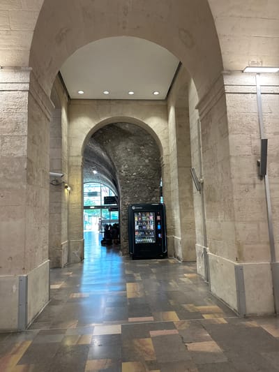 The image depicts an interior passageway with arched stone architecture. In the foreground, the floor is tiled with large, irregularly shaped tiles in various shades of gray, yellow, and red. On the right side of the passageway, a black vending machine labeled "selecta" is positioned against a stone wall. Further down the passageway, a tunnel-like structure with a rough stone ceiling is visible, leading to an area with natural light, where several people are seated or standing. Two security cameras are mounted on the stone pillars framing the passageway. A modern light fixture is attached to a pole on the right side. The overall impression is that of an indoor public space, possibly a train station or a historical building with modern amenities. This scene is located within the Arènes de Nîmes, a Roman amphitheater in Nîmes, France, which now serves as a public venue.