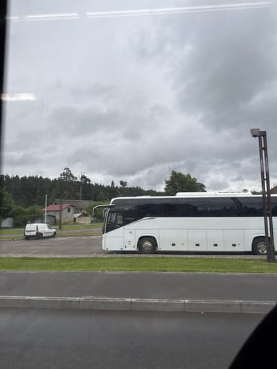 In the city of Rassudovo, Russia, a white bus with a black tinted window is parked in a lot. The bus is long and has multiple windows. In the background, there are trees and buildings. There is also a white van parked in the lot. The sky is cloudy and the ground is wet, suggesting that it may have rained recently. A lamppost is visible on the right side of the image.