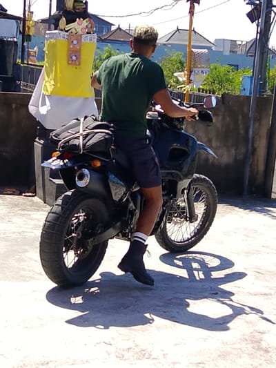 A person is on a black motorcycle, facing away from the camera, with a yellow and white object on the back of the motorcycle. The person is wearing a green t-shirt and dark shorts, with black boots and white-striped socks. The motorcycle has a black bag strapped to the back. The scene appears to be in an outdoor area with a concrete surface and a wall in the background. Buildings and trees are visible in the distance. The shadow of the motorcycle and the person are cast on the concrete surface. The image is taken on a sunny day. Based on the architecture of the buildings and the style of the object on the motorcycle, this location is likely Tabanan, Indonesia.